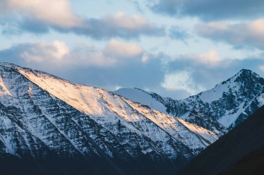 Atmosferik dağlık araziden, gün batımında karlı dağ sırtına. Dağ zirvesinde kar altın ışıkta parlar. Güzel, parlak, karlı bluz. Akşam bulutlu gökyüzü. Altın gün batımı gölgeleriyle harika bir manzara.