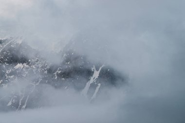 Alçak bulutların içinde karlı dağların olduğu manzaralı bir dağ manzarası. Yoğun siste güzel bir buzul. Bulutların arasından sabah ışığı. Bulutlu gökyüzünde kayalar ve pastel tonlarda hayalet atmosfer manzarası..