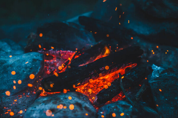 Vivid smoldered firewoods burned in fire closeup. Atmospheric background with orange flame of campfire. Wonderful full frame image of bonfire with glowing embers in air. Warm logs, bright sparks bokeh