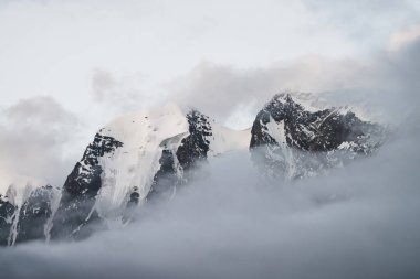 Alçak bulutların içinde karlı dağların olduğu manzaralı bir dağ manzarası. Yoğun siste güzel bir buzul. Bulutların arasından sabah ışığı. Bulutlu gökyüzünde kayalar ve pastel tonlarda hayalet atmosfer manzarası..