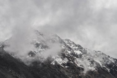 Atmosferik minimalist dağ manzarası ve karlı dağ zirvesi. Kar sınırına yakın alçak bulutlar. Karlı ve sisli kayalar. Sisli Craggy Dağları. Yüksek irtifada görkemli sisli manzara