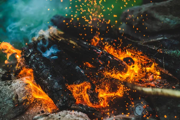 火の近くで焼かれた鮮やかな色付きの薪 キャンプファイアと青煙のオレンジ色の炎と大気中の背景 たき火の暖かいフルフレームイメージ 空気中のメンバーを育てる ボケの明るい火花 — ストック写真