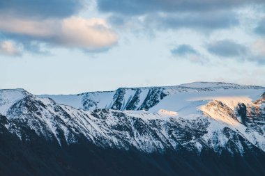 Atmosferik dağlık araziden, gün batımında karlı dağ sırtına. Dağ zirvesinde kar altın ışıkta parlar. Güzel, parlak, karlı bluz. Akşam bulutlu gökyüzü. Altın gün batımı gölgeleriyle harika bir manzara.