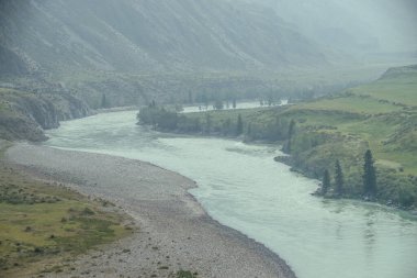 Geniş dağ nehri olan güzel sisli dağ manzarası. Koyu yeşil kasvetli manzara ve sisli dağ nehri. Yağmurlu havada, büyük dağların arasındaki büyük nehrin karanlık atmosferik görüntüsü.