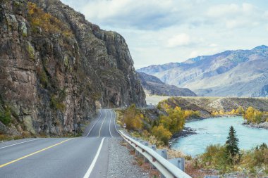 Parlak sonbahar manzarası, dağ yolu boyunca büyük dağ nehri boyunca güneş ışığı. Sonbahar renklerinde geniş turkuaz nehir ve dağ yolu olan parlak alp manzarası. Sonbahar zamanında dağlarda otoyol