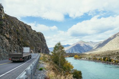 Sonbaharda gün ışığında dağlık nehir boyunca giden bir kamyon. Geniş turkuaz nehir ve dağ yolu. Dağlarda otoyol. Rusya, Altai Cumhuriyeti, Chuysky yolu, 19 Eylül 2020.