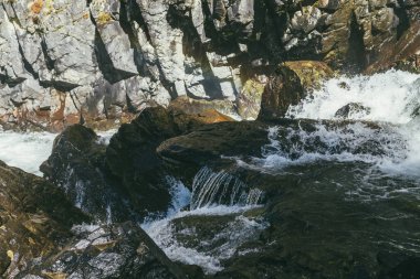 Atmosferik dağ manzarası, çalkantılı dağ nehri, kayalık duvarın yakınındaki yosunlu kayalar arasında güneş ışığı. Güneşin altında güçlü dağ nehrinde ıslak yosunlu taşlarla güzel bir dağ manzarası..