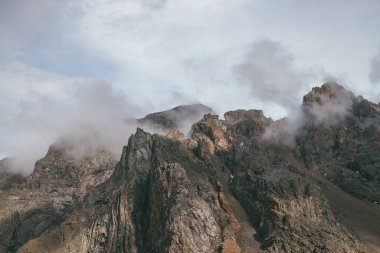 Görünüşte dağ manzarası, bulutlu gökyüzünde harika kayalar, klasik renklerde. Muhteşem güneşli manzara, alçak bulutlar kayalık dağın tepesinde pastel tonlarda. Güneş ışığı ve alçak bulutlarda güzel kahverengi kaya