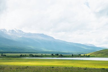 Dağ gölü ve ormanın olduğu dramatik bir manzara güneş ışığında tepeler ve değişken havada alçak bulutlarda karlı dağlar. Bulutlu gökyüzünde yüksek dağlar ve yeşil otların ve orman tepelerinin yakınında göl.