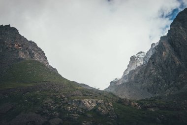 Yüksek kayalık dağlar ve alçak bulutlar arasında dar vadiyle atmosferik dağ manzarası. Bulutlu gökyüzünün altındaki güzel dağ vadisi. Keskin kayaların arasındaki vadiyle muhteşem bir manzara. Hayal edilemez görünüm.