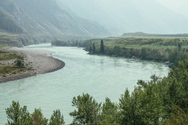 Geniş dağ nehri olan güzel sisli dağ manzarası. Koyu yeşil kasvetli manzara ve sisli dağ nehri. Yağmurlu havada, büyük dağların arasındaki büyük nehrin karanlık atmosferik görüntüsü.