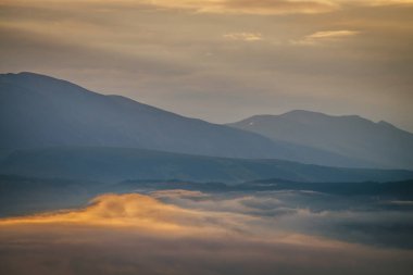 Bulutlu gökyüzünün altındaki mavi dağ siluetleri arasında vadide altın alçak bulutlarla şafak manzarası. Gün batımı ya da gün doğumu manzarası. Dağ vadisinde alçak bulutlar aydınlatıcı renkte..