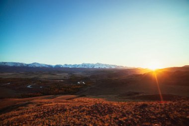 Orman vadisindeki dağ nehri ile muhteşem sonbahar manzarası ve altın güneş ışınlarıyla kaplı dağ sıraları. Tepeden karlı dağlara ve sonbahar vadisinden altın güneş ışınlarıyla muhteşem bir manzara.