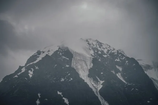 鉛灰色の曇りの空に黒い岩の上に氷河と暗い大気山の風景 雨天時の低雲の中に雪の山 霧の中で雪の黒い岩の山と悲観的な風景 — ストック写真