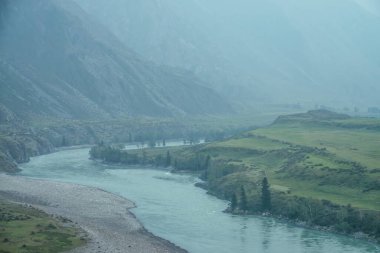 Geniş dağ nehri olan güzel sisli dağ manzarası. Koyu yeşil kasvetli manzara ve sisli dağ nehri. Yağmurlu havada, büyük dağların arasındaki büyük nehrin karanlık atmosferik görüntüsü.