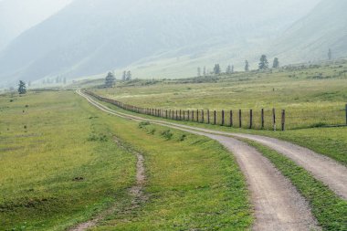 Güzel yeşil dağ manzarası çit boyunca uzun toprak bir yol ve sisli büyük dağlar. Atmosferik sisli dağ manzarası tepeler ve dağlar arasında toprak yol. Kırsalda uzun yol