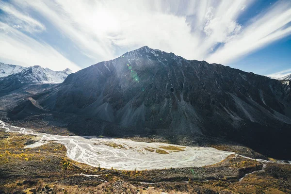 Beautiful alpine landscape with great sharp rocky peak and mountain river with many streams in valley in autumn color in sunshine. Awesome sunny scenery with high mountain wall with snow in sunlight.