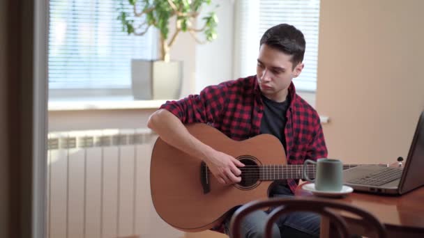 Un joven aprende a tocar la guitarra a través de Internet, portátil, lección en línea. Hobby y concepto de ocio — Vídeo de stock