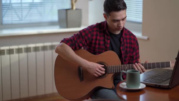 Un joven aprende a tocar la guitarra a través de Internet, portátil, lección en línea. Hobby y concepto de ocio — Vídeo de stock