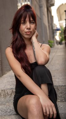 Şık bir kadın, kızıl saçlı ve dövmeli, şehir merdivenlerinde oturuyor, kendine güvenen bir ifadeyle kameraya bakıyor.