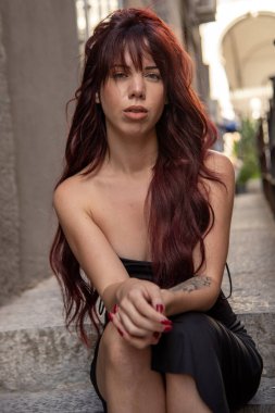 Long-haired redhead in a black dress sits on city stairs, gazing confidently at the camera with an elegant, urban pose