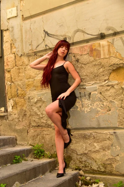 Siyah bir elbise ve yüksek topuklu ayakkabı giyen genç kızıl saçlı kadın eski bir duvar yazısı ve merdivenleri olan grunge şehir duvarına karşı modellik yapıyor.