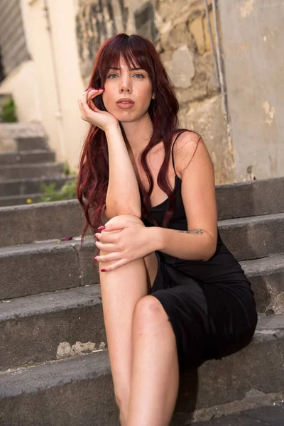 Siyah elbiseli kızıl saçlı kadın yıpranmış taş basamaklarda oturuyor, yoğun ve düşünceli bir ifadeyle kameraya bakıyor.