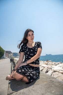 Duvarda oturan genç bir kadın, kameraya bakıyor, arka planda deniz ve şehir gezinti alanı var.