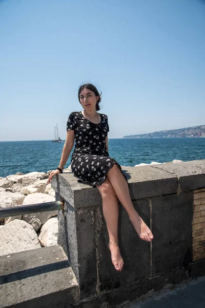 Genç bir kadın güneşli bir yaz gününde berrak mavi gökyüzünün altında sakin denizi gören tarihi bir taş duvarda yalınayak oturuyor.