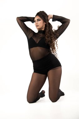 Beyaz arka planda diz çökmüş siyah bluz, şort ve tayt giymiş genç bir kadın. Anlamlı bir duruşla başka yere bakıyorsun.