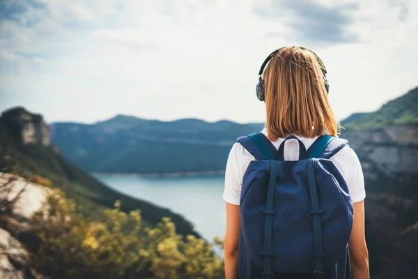 back view girl backpacker with light hair dreams listening to music in headphones relaxation on top rocky mountains, blonde hipster with backpack leisure after walking lake valley or green summer hills, empty space