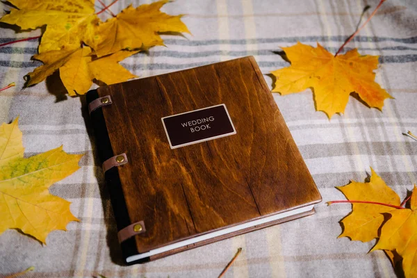 A leather-bound book with a designer wooden cover and a chocolate-colored tag lies on a tablecloth among yellow autumn leaves