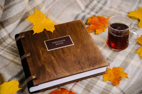 Leather-bound book with designer wood cover and chocolate-colored tag. The book lies on a gray blanket. next to a book a glass of tea and yellow maple leaves