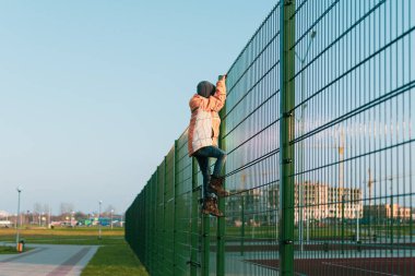 Kalın elbiseli küçük bir çocuk spor sahasının metal çitlerine tırmanıyor.