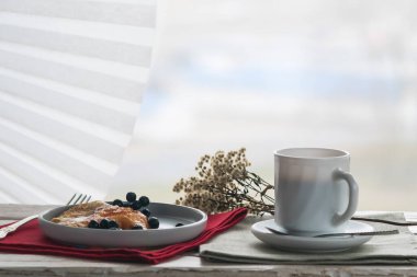 Dekoratif bir masanın üzerinde bir fincan kahve ve bir tabak krep, perdeli pencerenin arka planına karşı.