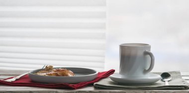 Dekoratif bir masanın üzerinde bir fincan kahve ve bir tabak krep, perdeli pencerenin arka planına karşı.