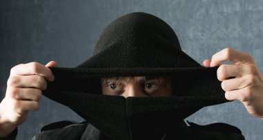 Kar maskesi takmış kahverengi gözlü genç bir adam karanlık bir arka planda kameranın önünde poz veriyor.