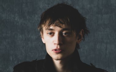 Kahverengi gözlü genç bir adam kameranın önünde koyu bir arka planda poz veriyor.