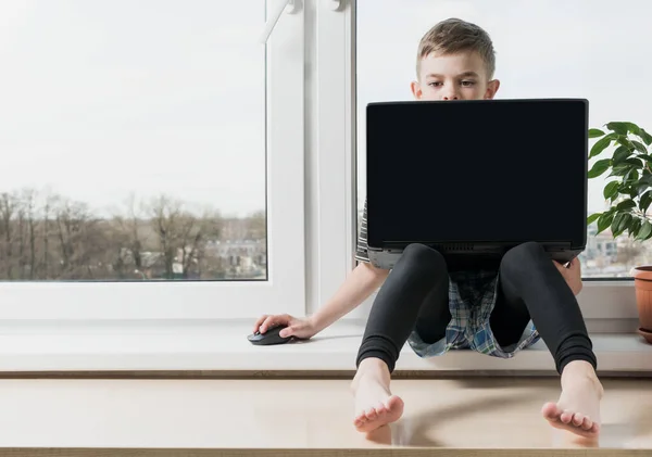 A small boy sits at a computer near the window in the apartment