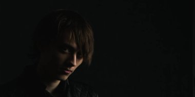 Çok koyu bir arka planda uzun saçlı bir gencin portresi.