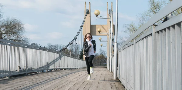 一个戴着时髦眼镜和皮夹克的小女孩在一座悬吊的人行天桥上奔跑 — 图库照片