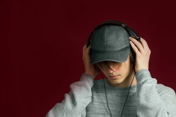 A young guy with long hair listens to music with headphones on a red background
