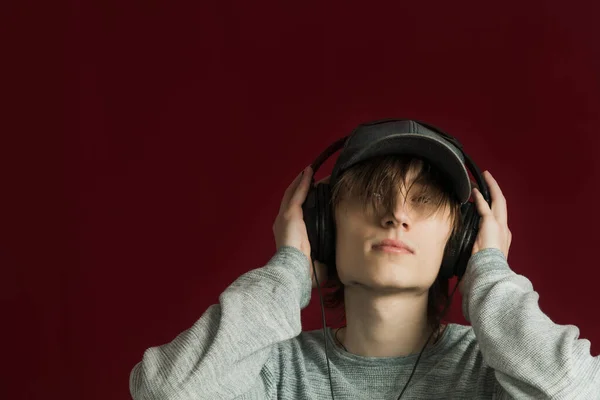 A young guy with long hair listens to music with headphones on a red background