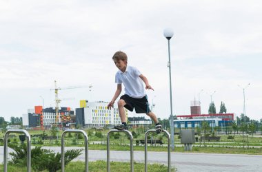 Beyaz tişörtlü küçük bir çocuk parkta oynuyor, metal korkuluklarda zıplıyor.