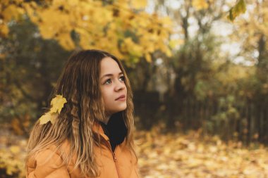 Sarı ceketli genç bir kız düşen yapraklarla bir sonbahar parkında yürüyor.