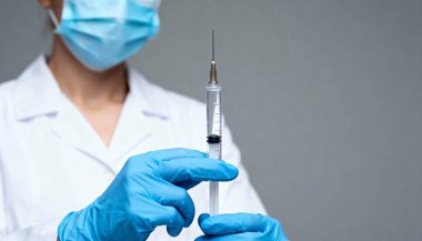 Beyaz önlüklü ve elinde şırınga tutan mavi cerrahi maskeli bir adam. Kişinin yüzü görünürde yok. Kişinin elleri mavi nitril eldivenlerle kaplıdır.