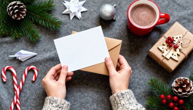 Noel kartı tutan bir çift el. Ellerin etrafına çeşitli Noel temalı dekoratif öğeler saçılmıştır: çizgili şeker kamışı, kağıt süslemeler, gümüş bir Noel süsü ve kozalaklı yeşil çam dalları..