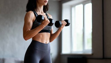 Yaklaşık 25 yaşında, bronz tenli, koyu renk saçlı, spor bir bluz ve tayt giyiyor. Yüzü görünmüyor, vücuduna odaklanmış durumda. Modern bir odada elinde dambıllarla dikiliyor.. 