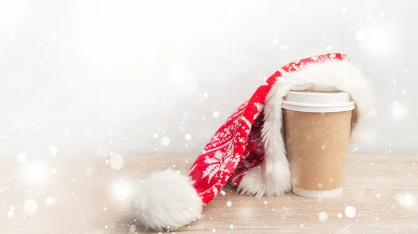 Banner xícara de café embrulhado em um chapéu de Papai Noel em um fundo de Natal branco e de madeira com neve — Fotografia de Stock