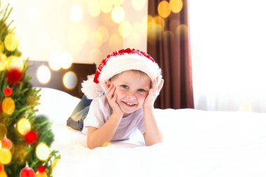 Noel Baba şapkalı çocuk Noel ağacının yanındaki yatakta yatıyor ve hediye bekliyor. Yeni yıl tebrik kartı